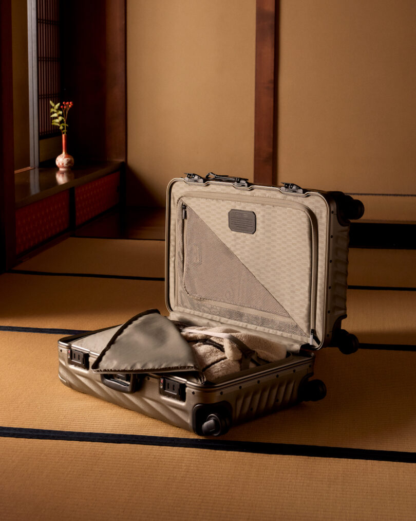 A beige suitcase is open on a tatami mat in a room with wooden walls and a small flower vase on a shelf