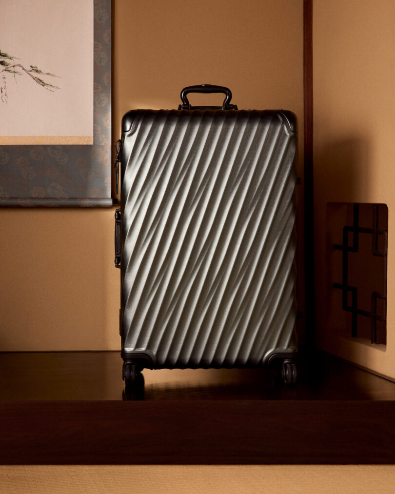 A textured silver suitcase stands upright on four wheels in a room with beige walls and a decorative framed picture