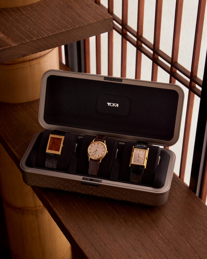 Three wristwatches with leather straps are displayed in an open black case with a logo, placed on a wooden surface near vertical blinds