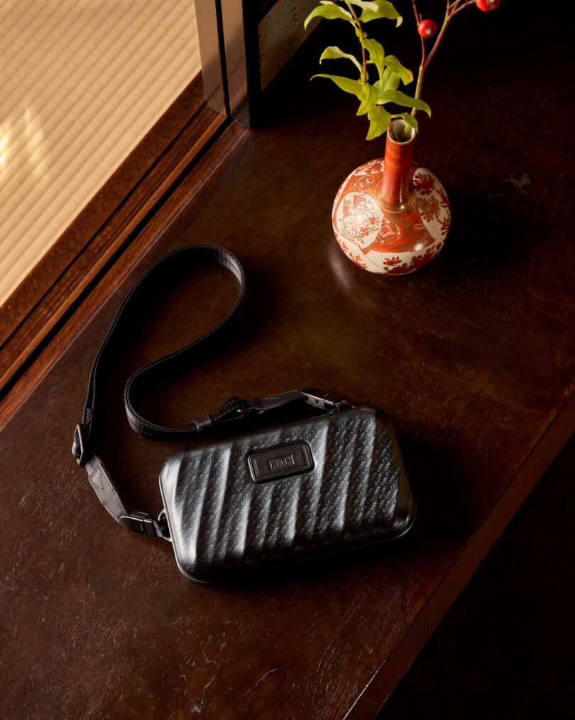 A black leather wristlet with a textured design rests on a dark wooden table next to a decorative vase with green leaves and red berries