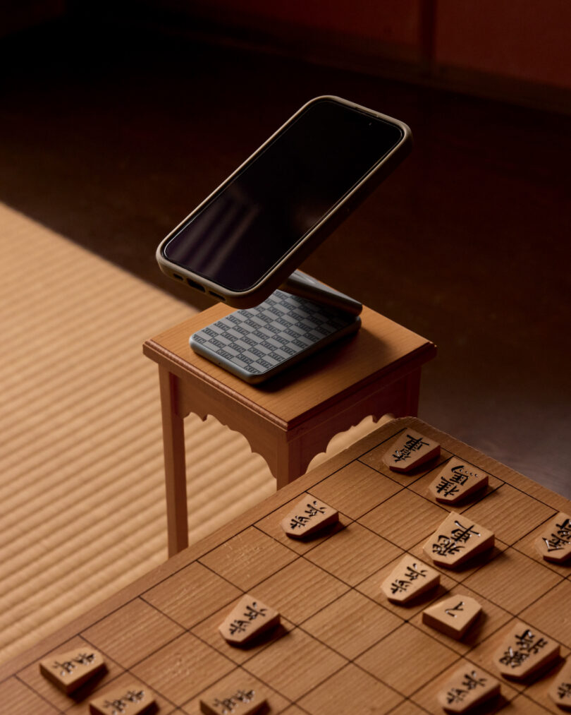 A smartphone on a stand is positioned on a small wooden table next to a shogi board with game pieces arranged on a tatami mat floor