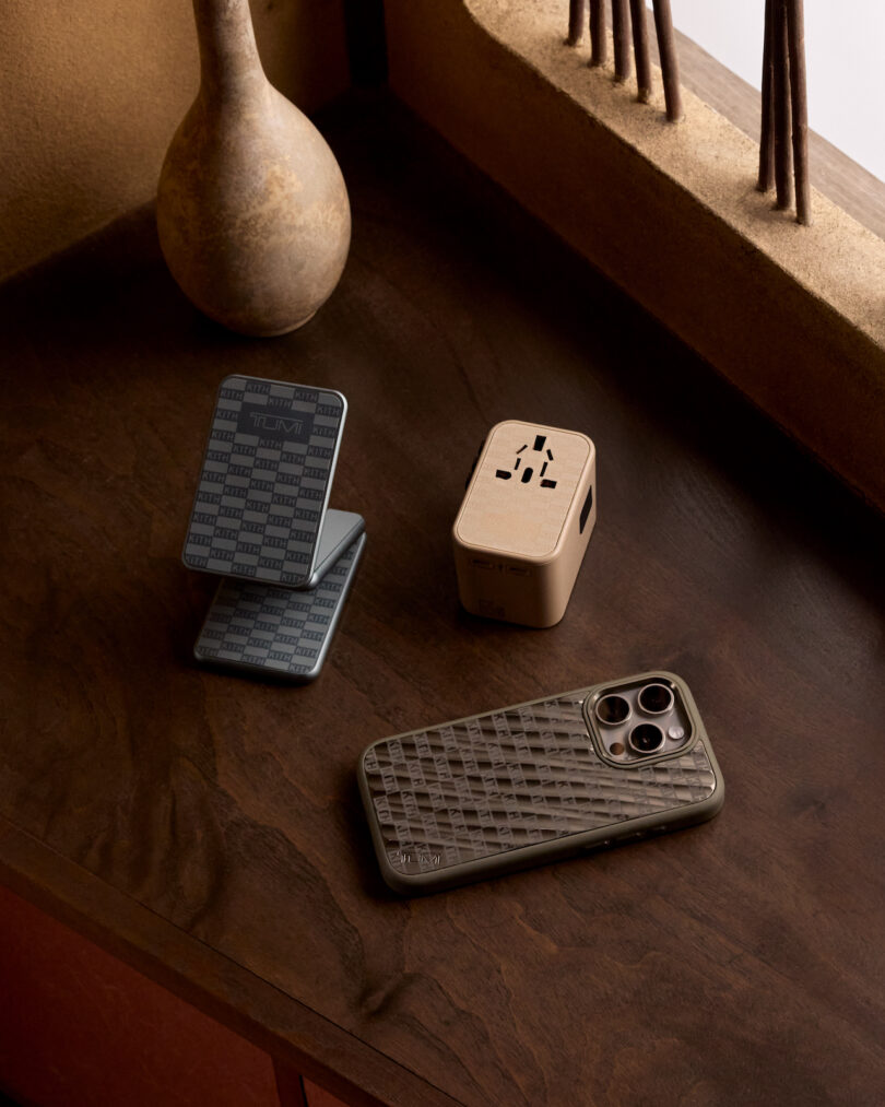 Smartphone with a textured case, a travel adapter, and a collapsible phone stand on a wooden surface with decorative vases in the background