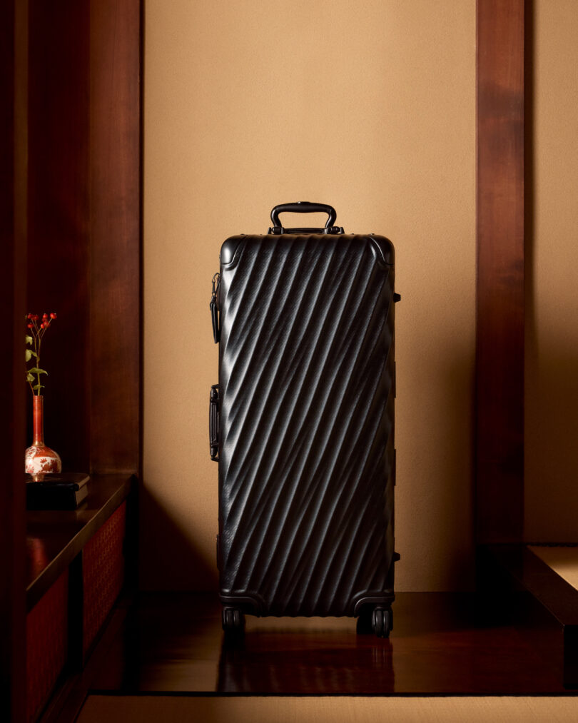 A black textured suitcase with four wheels stands upright in a warmly lit room with wooden accents. A small vase with red flowers is on a shelf to the left