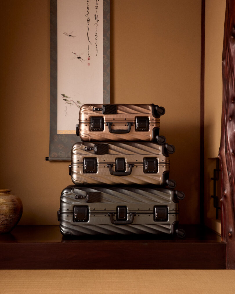 Three stacked suitcases in different shades of brown and gold sit on a wooden shelf against a beige wall, with a scroll and a ceramic vase nearby