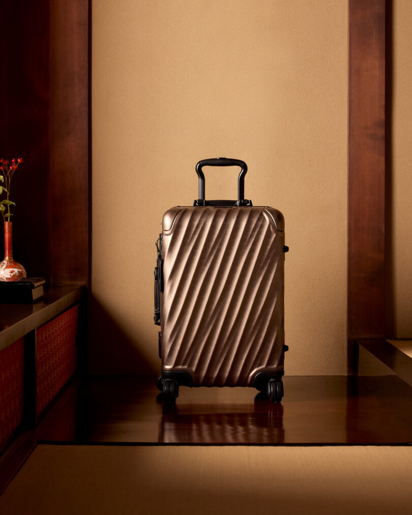 A metallic rolling suitcase stands upright on a wooden floor, flanked by a wall with a vase on the left