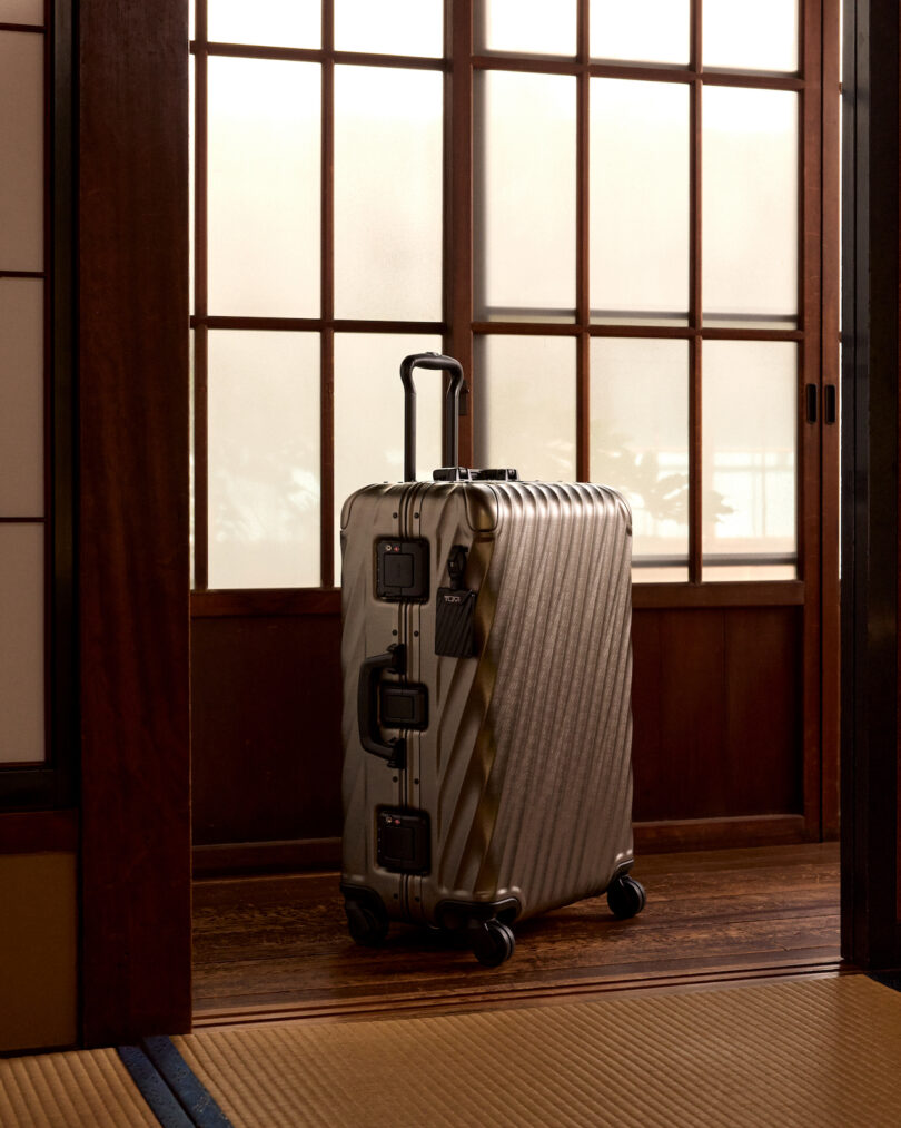 Silver suitcase with a striped design stands upright on wheels in front of a wooden shoji door. The room features tatami flooring