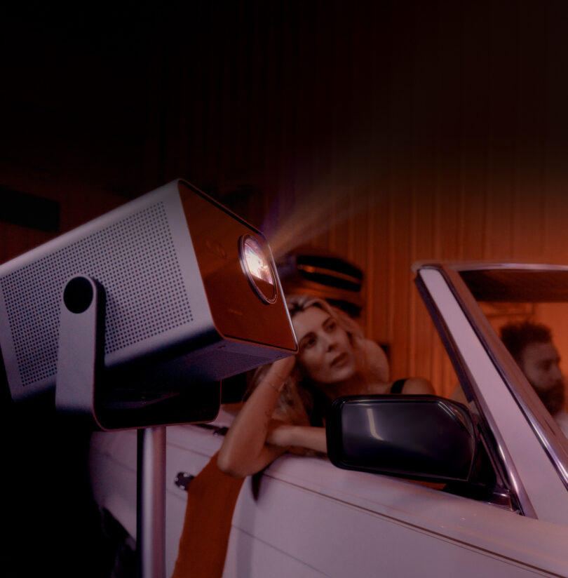 A projector casts light with a woman leaning on a convertible car in a dimly lit room.