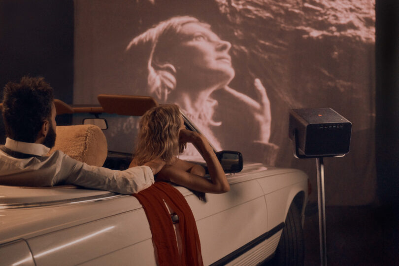 Two people sit in a convertible indoors, watching a black-and-white film projected on the wall. A vintage projector stands nearby.