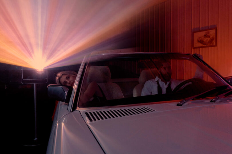 A man and woman sit in a convertible car, illuminated by colorful lights from a projector in a dimly lit room.