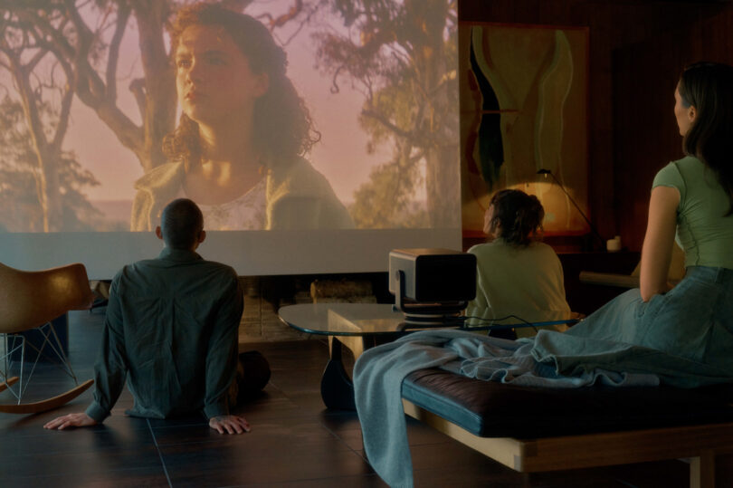 Three people watching a movie projected on a screen in a dimly lit room.