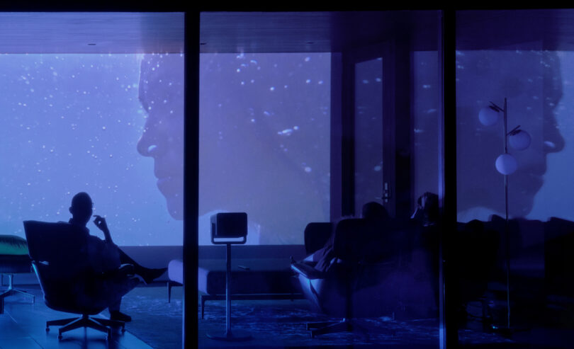 Silhouetted person sits in a dark room with two large projected faces on a blue-tinged screen, creating a contemplative atmosphere.