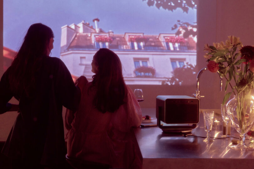 Two people stand in a dimly lit kitchen, facing a projected image of a building. A vase of flowers and a digital device are on the counter.