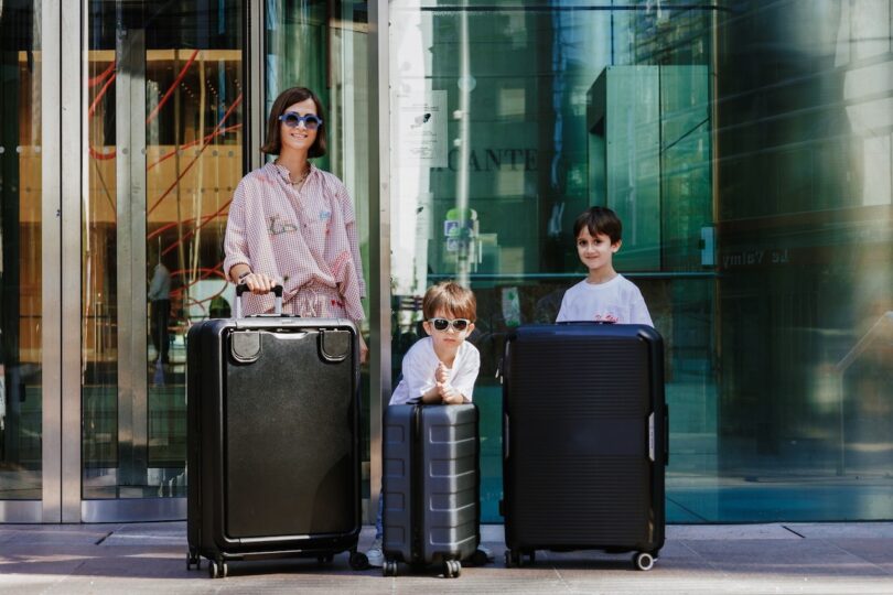 A woman with two children, all wearing sunglasses, stand with large black suitcases outside a building with glass doors