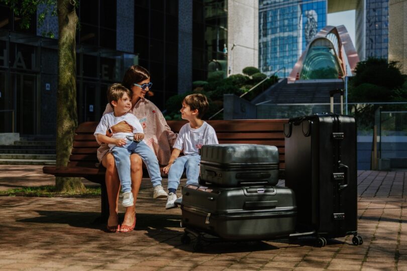 A woman sits on a bench with two children, surrounded by several large suitcases. They are in an outdoor urban setting with modern buildings in the background