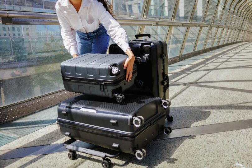 Person placing a black suitcase on top of two other black suitcases in a sunlit atrium with glass and metal structure