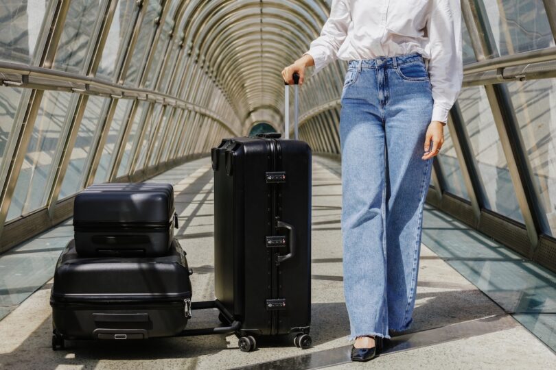 A person in a white shirt and blue jeans pulls a black suitcase through a glass-covered walkway. Two additional black bags are stacked on the suitcase