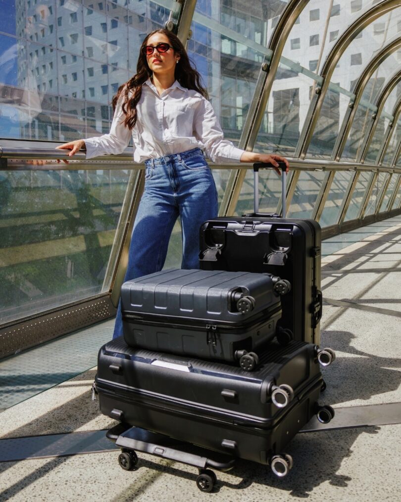 A woman in a white shirt and jeans stands with three stacked suitcases in a glass bridge or hallway