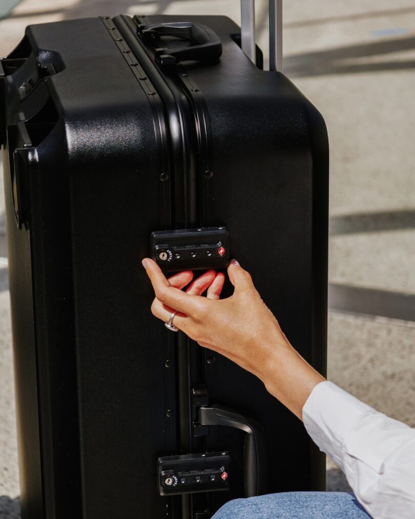 A person with red nail polish adjusts a black suitcase handle on a sunny day