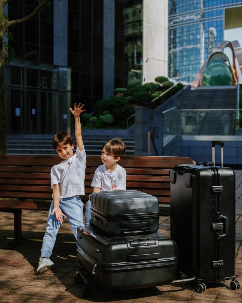 Two children sit on a bench next to stacked suitcases in an urban setting