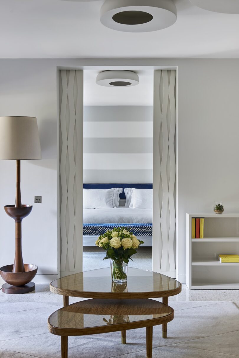 A modern living room with a round wooden table, a lamp, and a bookshelf. A bouquet of yellow roses is on the table. A doorway reveals a bed with striped bedding in the adjacent room.