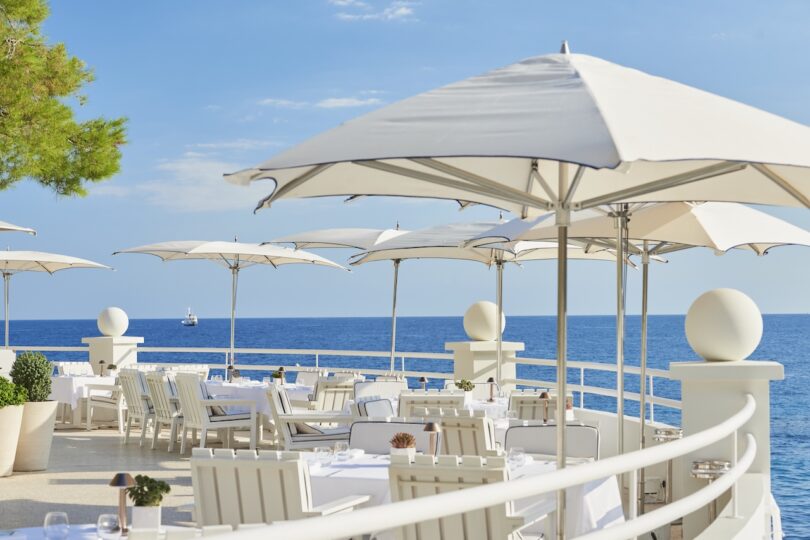 Outdoor dining area with white tables, chairs, and umbrellas overlooking a calm ocean. A small boat is visible in the distance under a clear blue sky.