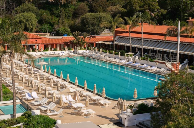 Outdoor swimming pool surrounded by lounge chairs and umbrellas, with terracotta-roofed buildings and lush greenery in the background.