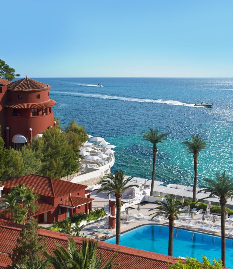 Oceanfront view with red-roofed buildings, a pool, palm trees, and a boat on the blue sea.