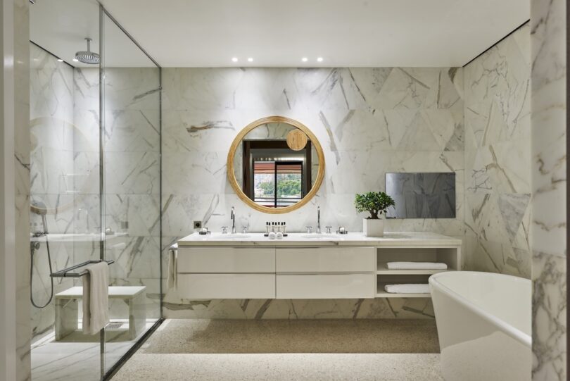 Modern bathroom with marble walls, a round mirror, double sink vanity, glass shower, and freestanding bathtub. A small plant decorates the counter.