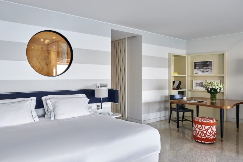 Modern hotel room with a striped wall, round mirror, bed with white linens, desk with a chair, floral arrangement, and red-patterned stool.