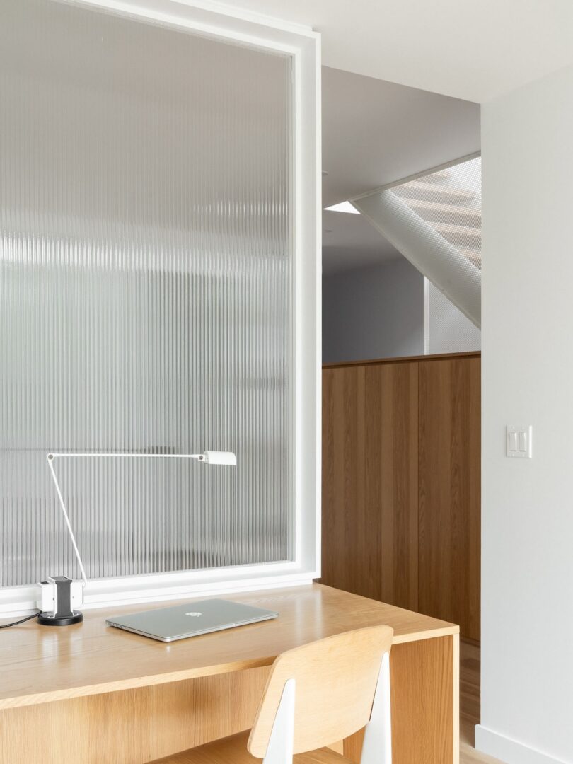 A minimal workspace features a wooden desk, a modern lamp, and a closed laptop. A frosted glass panel and light wood paneling are visible in the background.