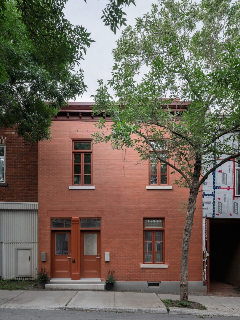 Two-story brick house with brown doors and windows, flanked by a tree on the right and neighboring buildings.