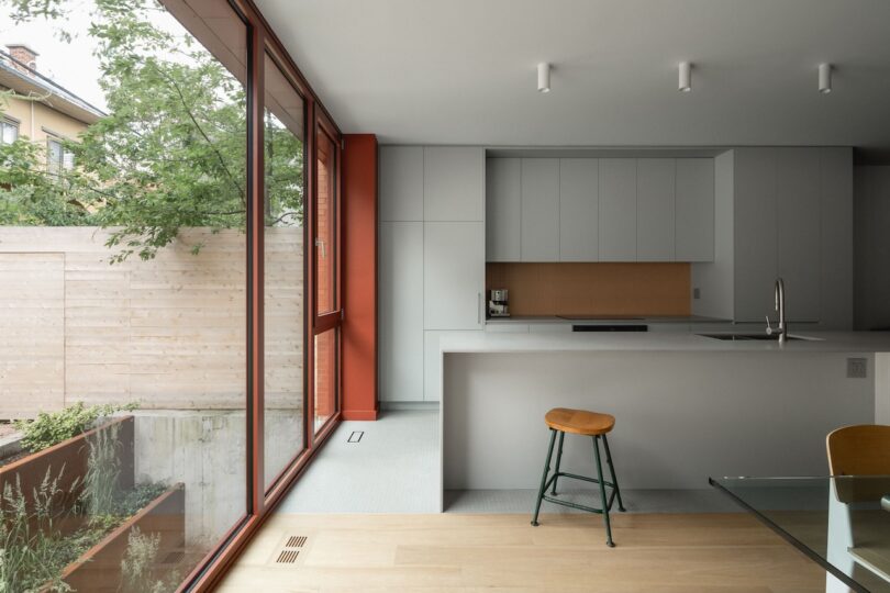 Modern kitchen with large windows, white cabinets, and a central island with a stool. Glass table in the foreground and view of a wooden fence outside.