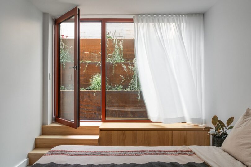 A bedroom with a wooden bed, white curtains, and an open door leading to a garden with plants.