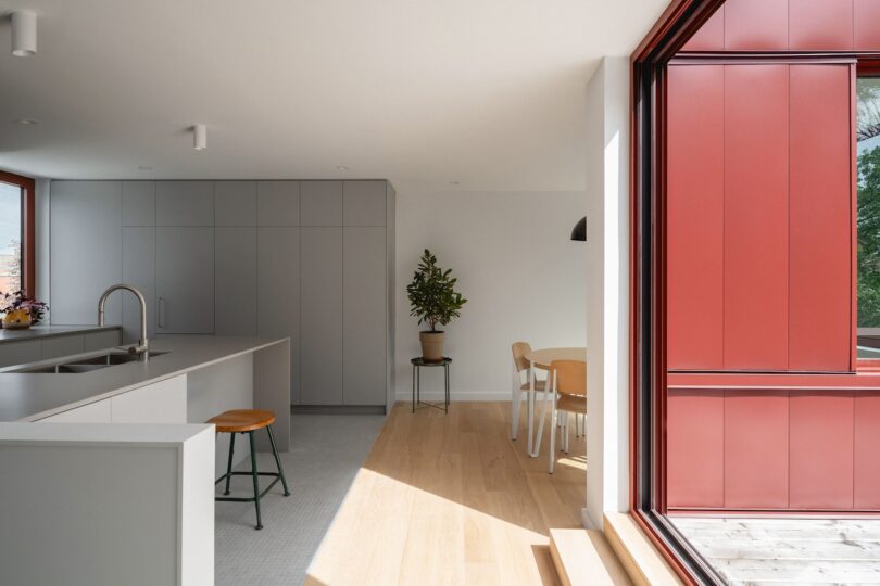 Modern kitchen with gray cabinets, an island with a sink, and a stool. Adjacent is a dining table with chairs. Large red-framed windows allow natural light to fill the space.