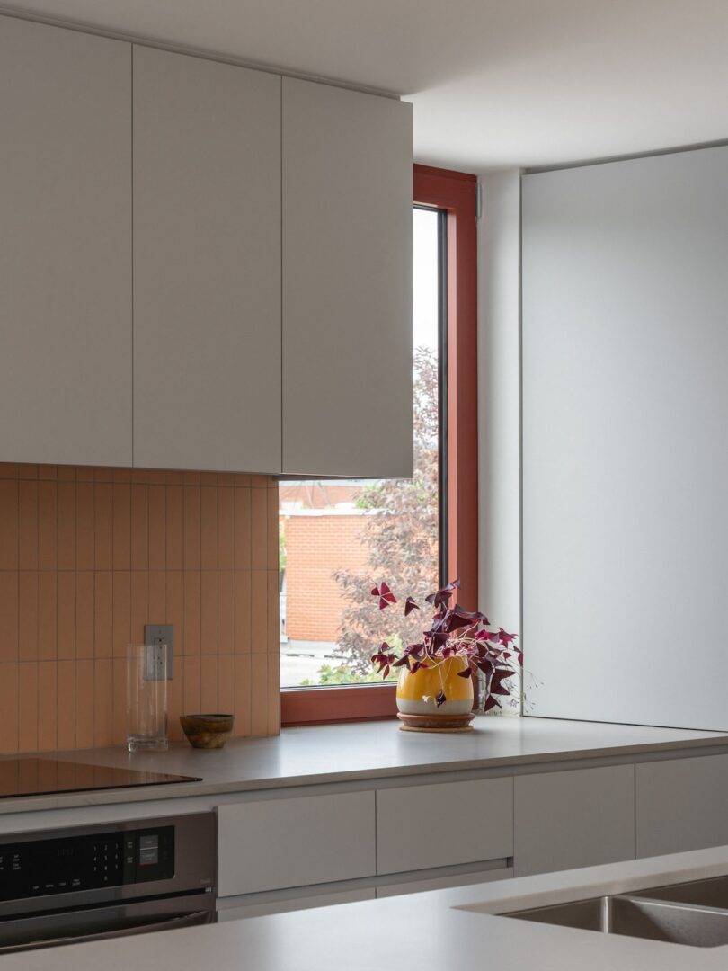 Modern kitchen with white cabinets, beige tile backsplash, and a stainless steel sink. A window provides natural light. A yellow pot with a potted plant sits on the countertop.