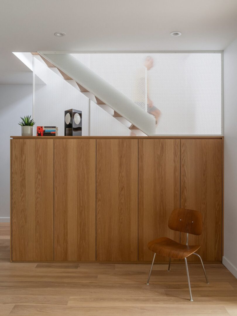 Wooden cabinet and chair beneath a staircase with a mesh screen. A small plant, books, and a clock sit atop the cabinet.