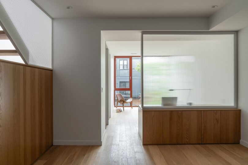 Modern interior with wooden floors, white walls, and large frosted glass panel. A corridor leads to a room with a wooden chair and a large window providing natural light.