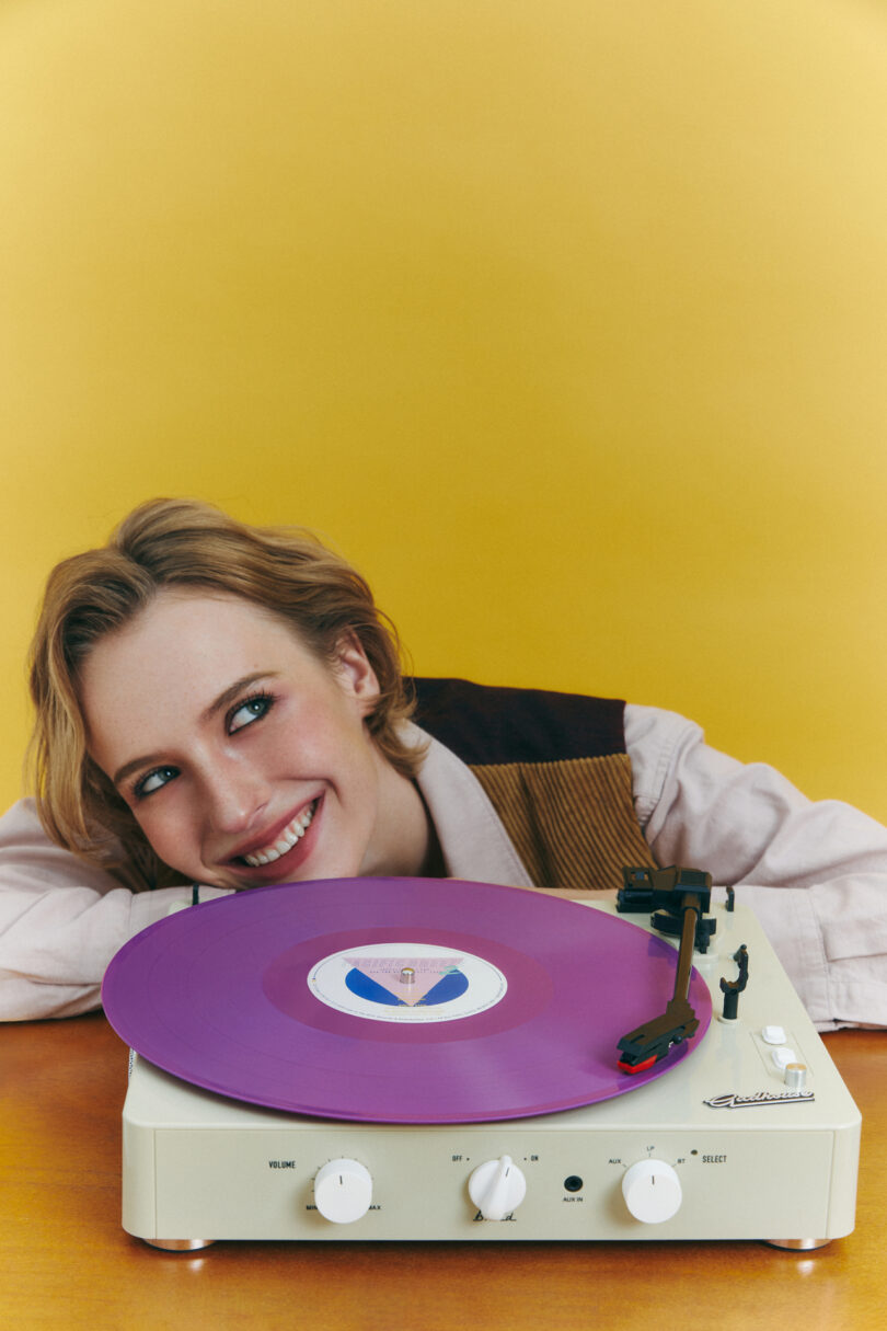 A person smiles while resting their head on a table next to a record player with a purple vinyl against a yellow background.