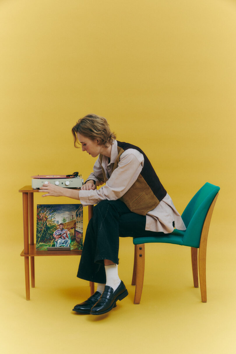 A person sits on a chair with teal upholstery, operating a vintage typewriter on a small wooden table against a mustard yellow background. A colorful painting leans against the table.