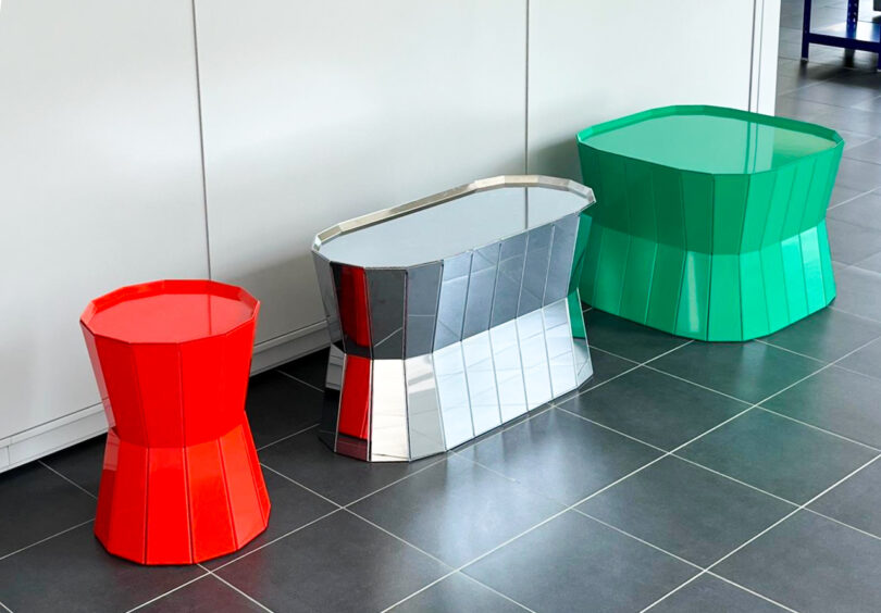 Three geometric, colorful stools: one red, one silver with red and black sections, and one green, placed on a tiled floor in front of a white wall