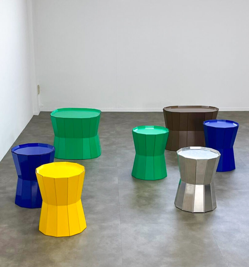 Several colorful, geometric stools are arranged on a gray floor in a minimalist room with white walls