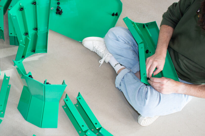 Person sitting on the floor assembling green metal panels, wearing a green hoodie, light jeans, and white sneakers