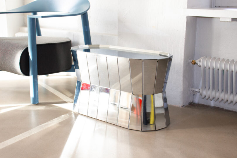 A modern metal vase with a reflective surface is placed on the floor near a blue chair and a radiator