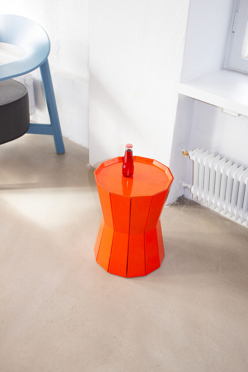 A red octagonal stool with a small red bottle on top, placed near a radiator and a blue chair against a white wall