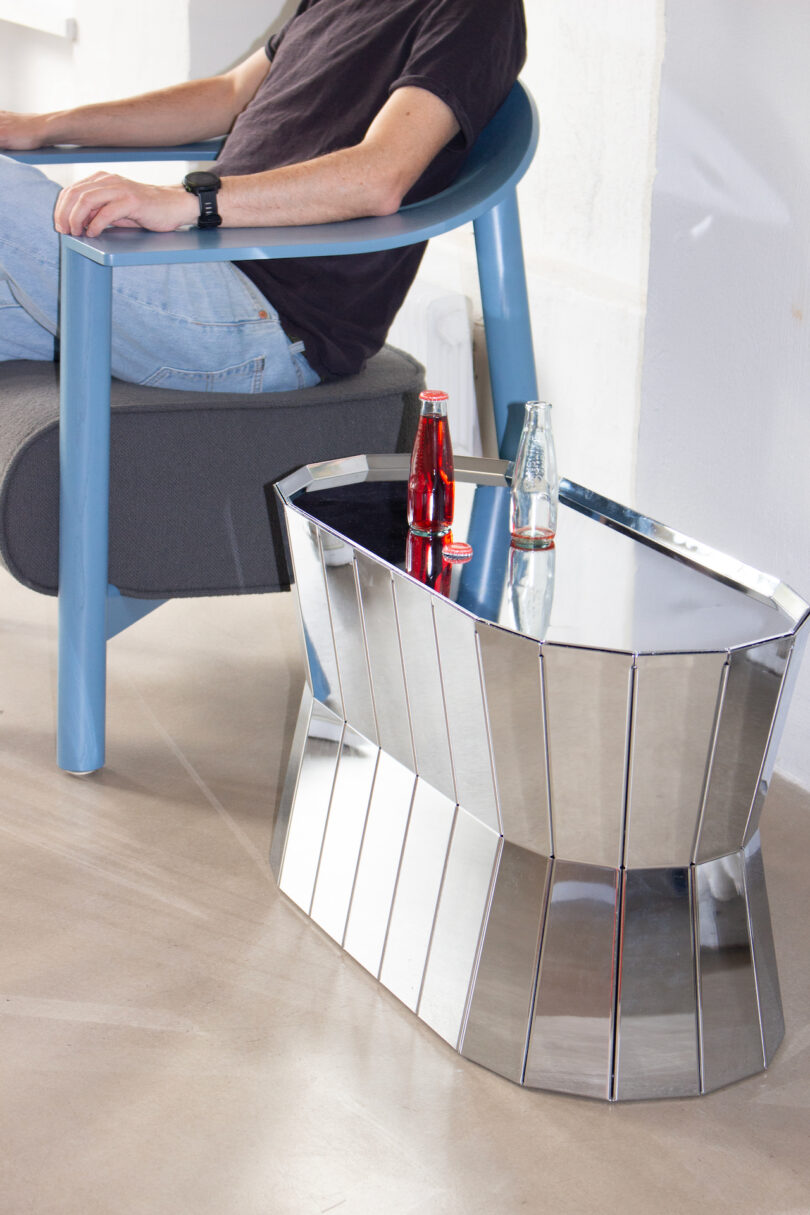 Person seated in a blue chair near a modern, metallic side table with a red soda bottle, a glass, and a bottle cap on it