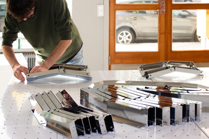 A person assembles metal components on a large workstation, with several metal pieces laid out on the table. A car is visible through the window in the background