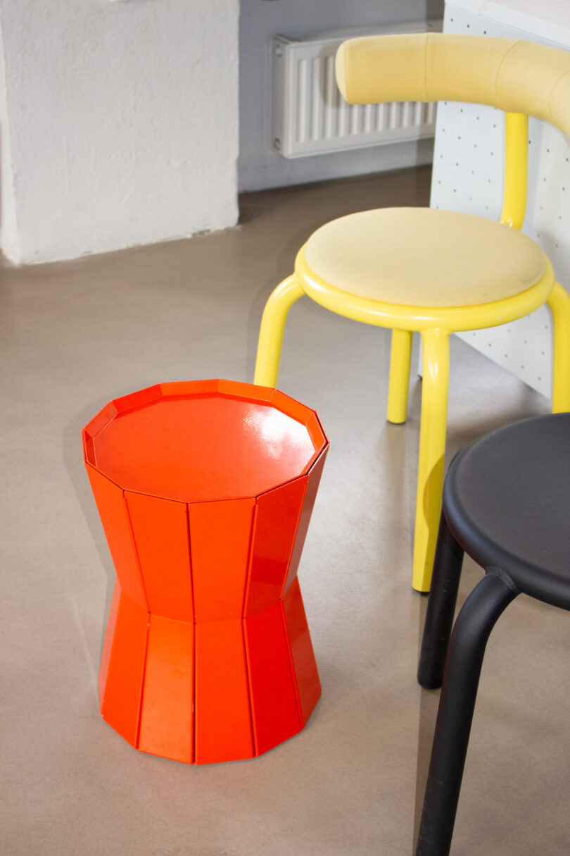 A bright orange stool next to a yellow chair and a black seat on a beige floor