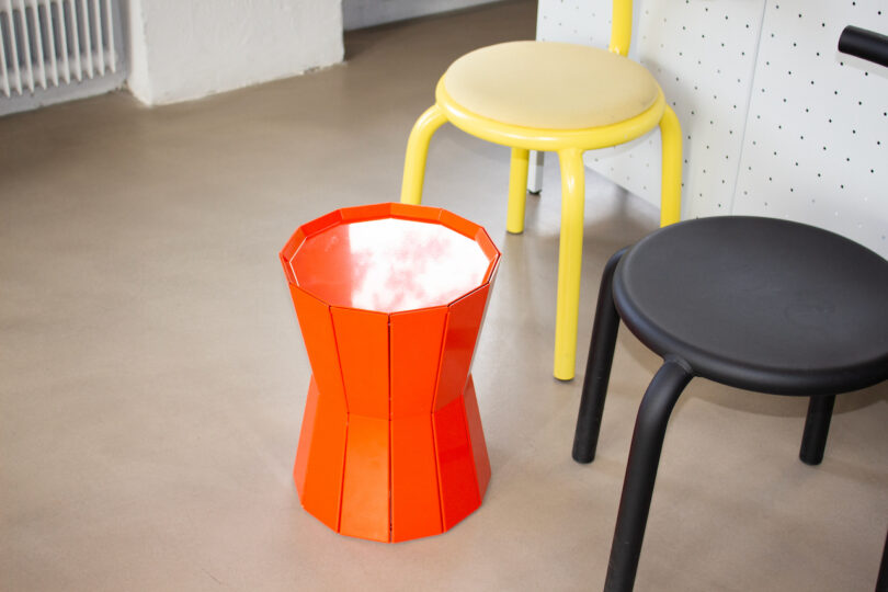 Three stools are placed on a beige floor: an orange geometric stool, a round yellow stool, and a round black stool