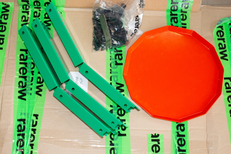 Green curved metal pieces, an orange plastic disc, and a bag of screws are displayed on a cardboard surface marked "rareraw"