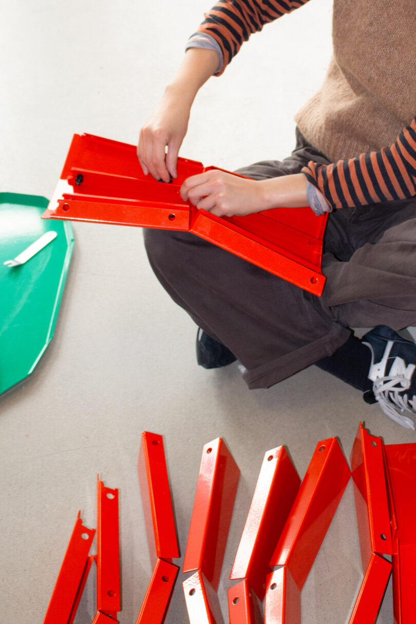 Person assembling red metal parts on the floor, wearing a striped long-sleeve shirt and sneakers